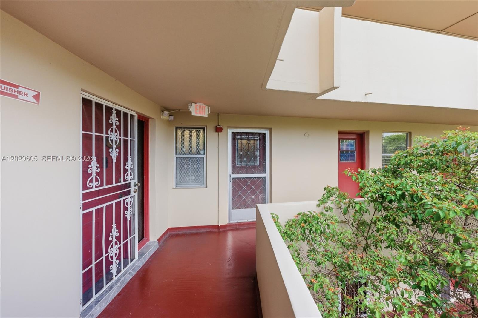 An outdoor apartment walkway features red doors, security gates, and a view of greenery — 1501 NE 191st St # C217, Miami FL 33179, MIAMI, FL