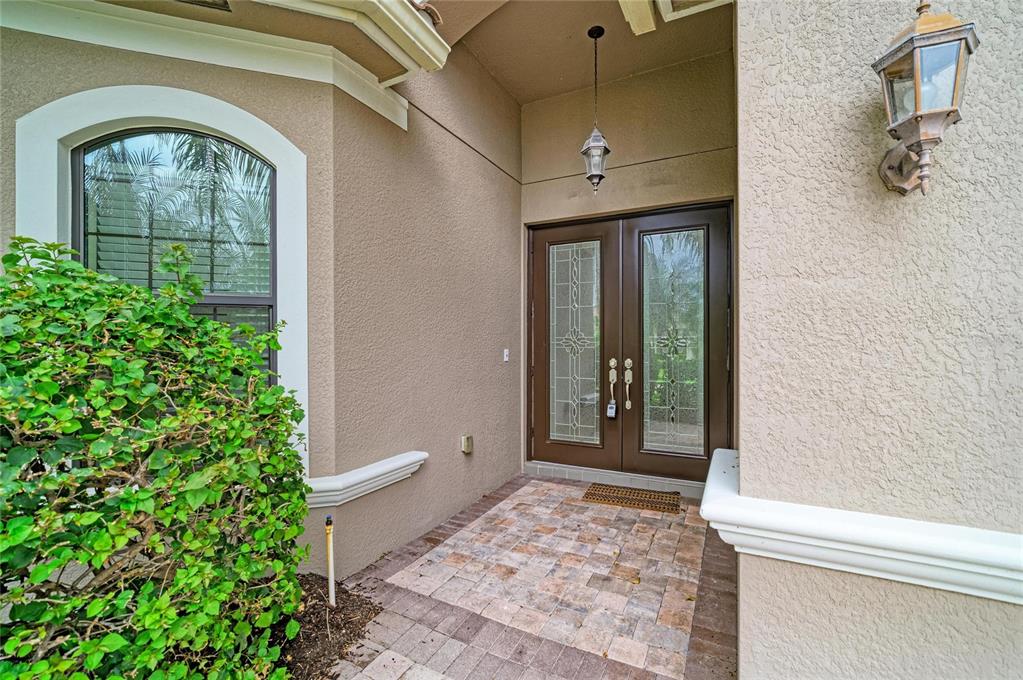 A beige stucco home entrance features a double glass door, stone pavers, and wall lighting — 3701 PLEASANT SPRINGS DRIVE, NAPLES, FL