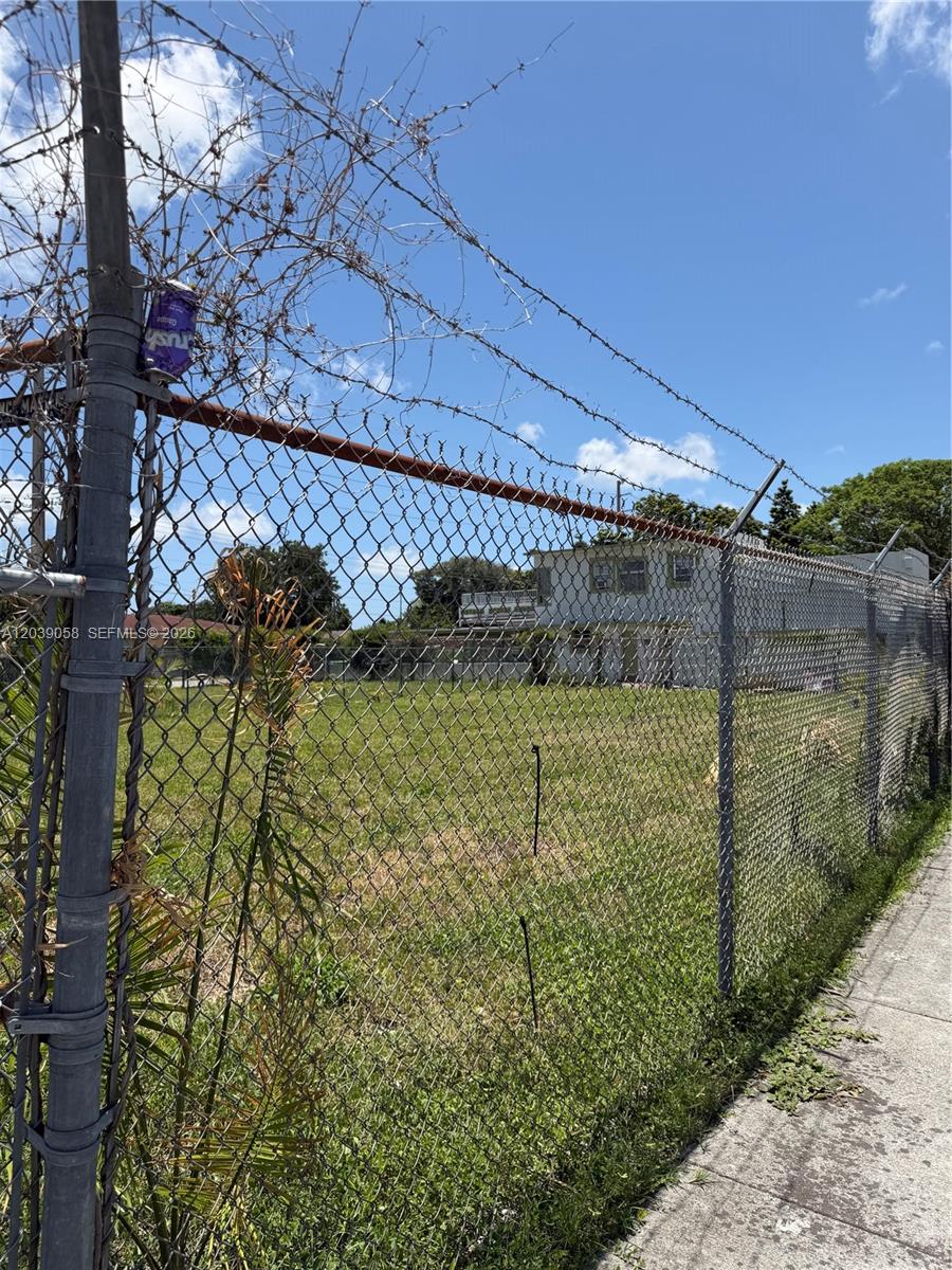 A chain-link fence with barbed wire surrounds a vacant, grassy lot under a blue sky — 810 NW 79th St, Miami FL 33150, MIAMI, FL