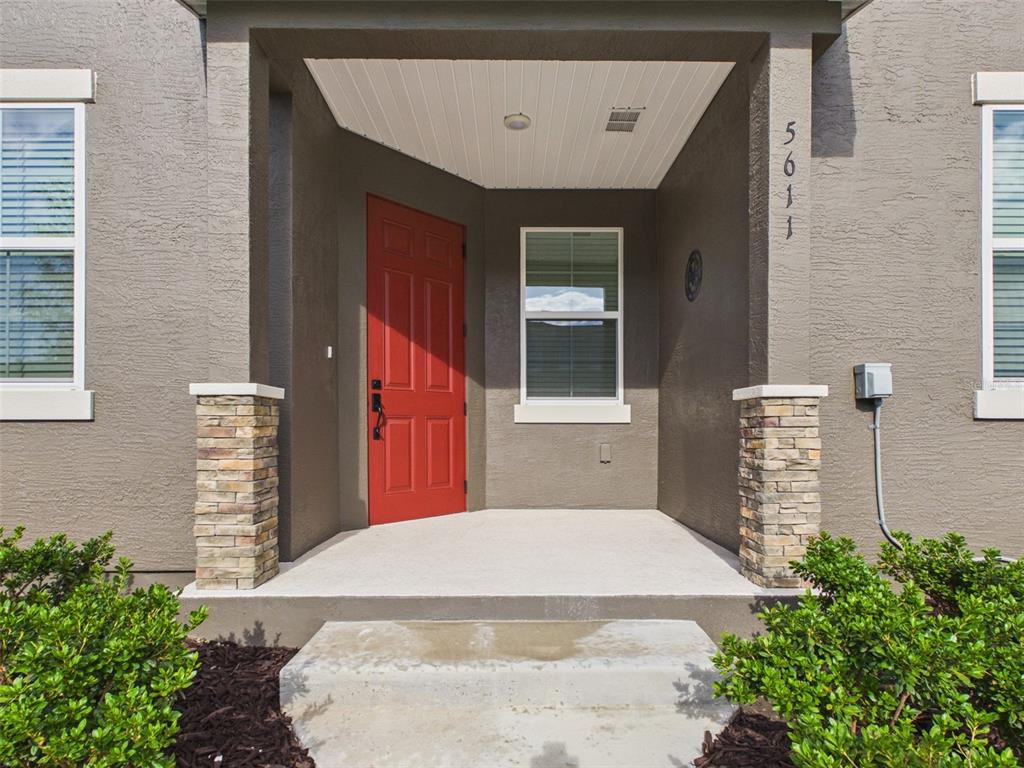 A covered concrete porch features a red front door, stone-accented columns, and exterior lighting — 5611 SW 72ND COURT, OCALA, FL