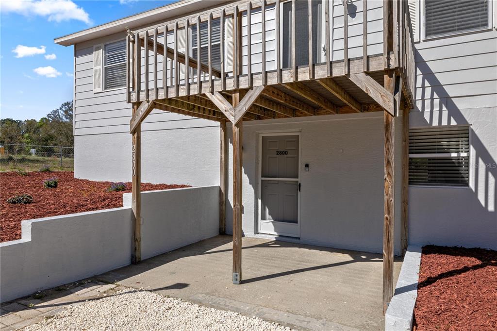 A ground-level concrete patio sits beneath a wooden balcony attached to a white house — 2800 SW 141ST COURT, OCALA, FL