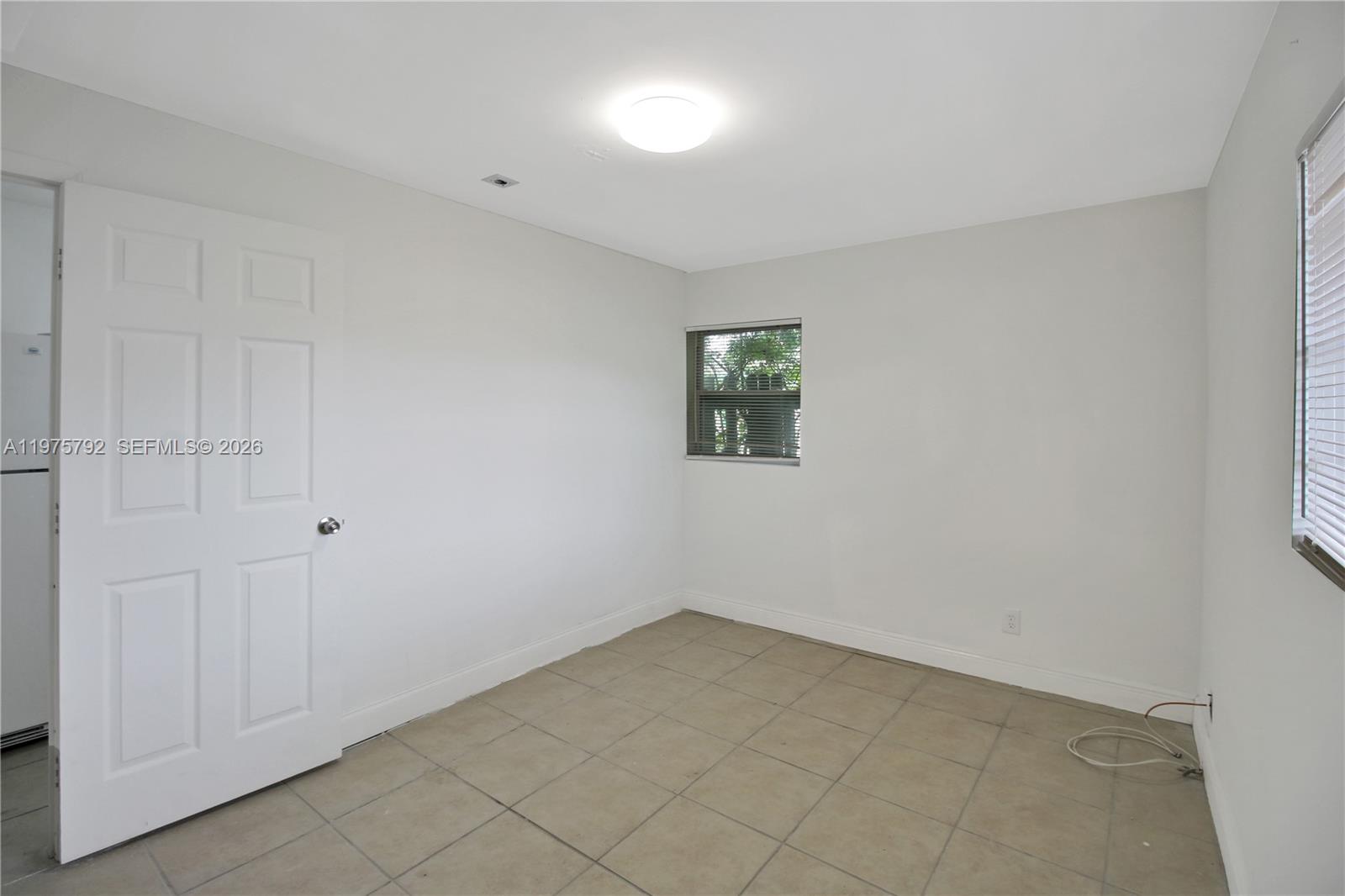 A small, empty room features white walls, tile flooring, a white door, and two windows — 1160 W 26th ct # D, Riviera Beach FL 33404, RIVIERA BEACH, FL