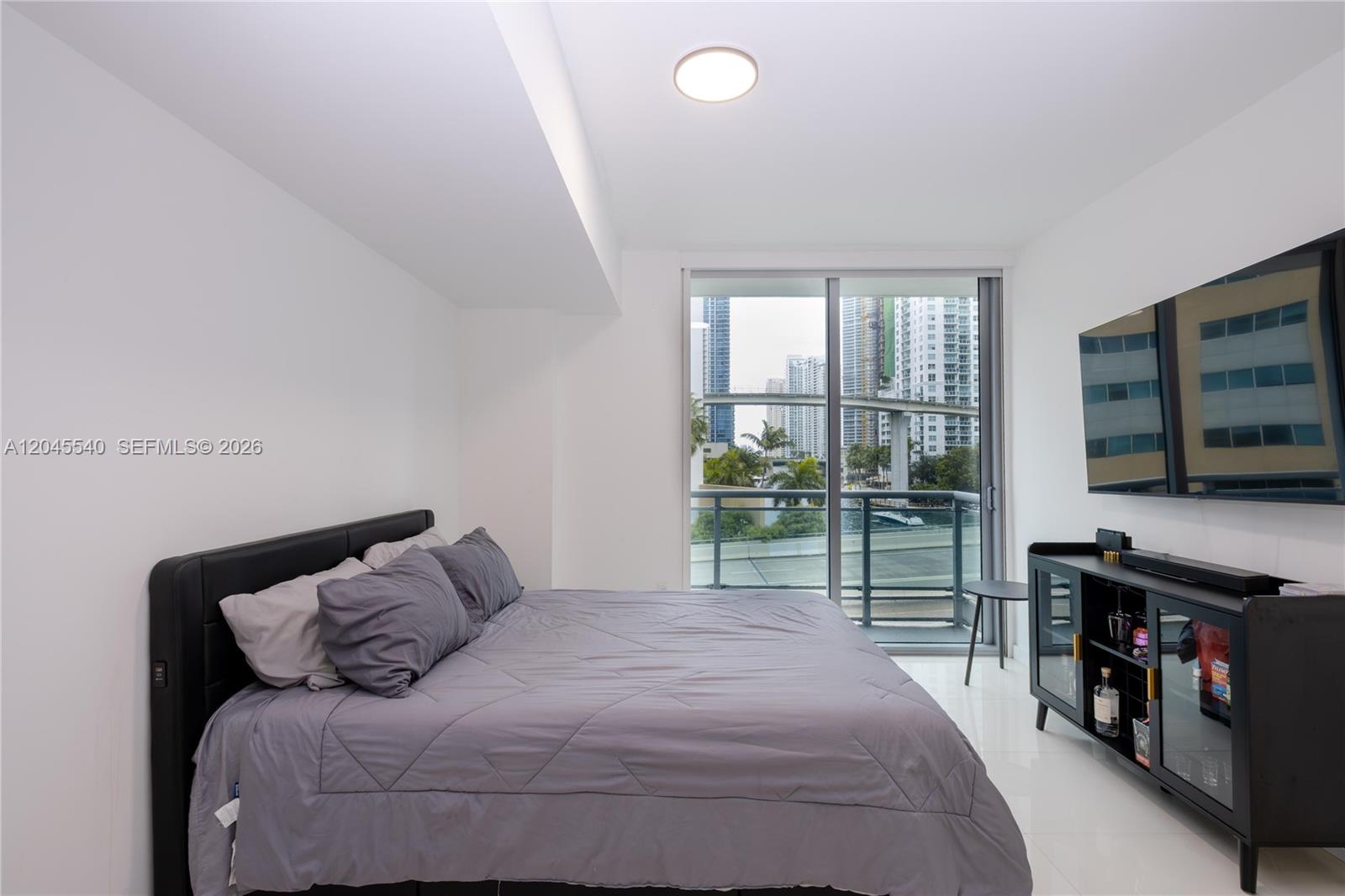 A bedroom features a bed, a wall-mounted television, and a balcony overlooking a city skyline — 350 S Miami Ave # 306, Miami FL 33130, MIAMI, FL