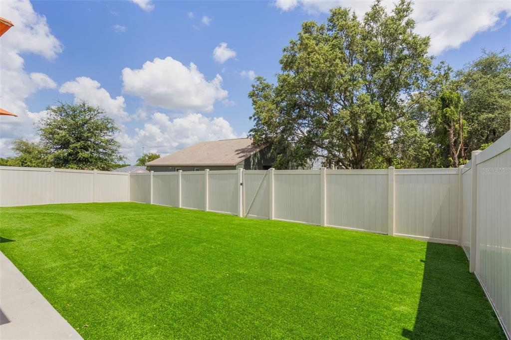A fenced backyard features artificial turf grass and a large tree under a blue sky — 840 TETON STREET, HAINES CITY, FL