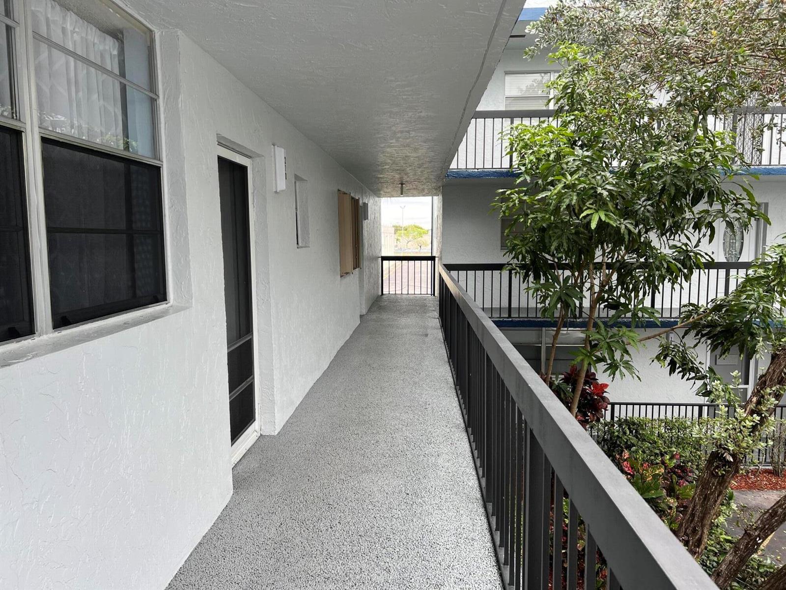 An outdoor apartment walkway features a gray textured floor, white walls, and a black railing — 3220 Holiday Springs Boulevard # 210, Margate FL 33063, MARGATE, FL
