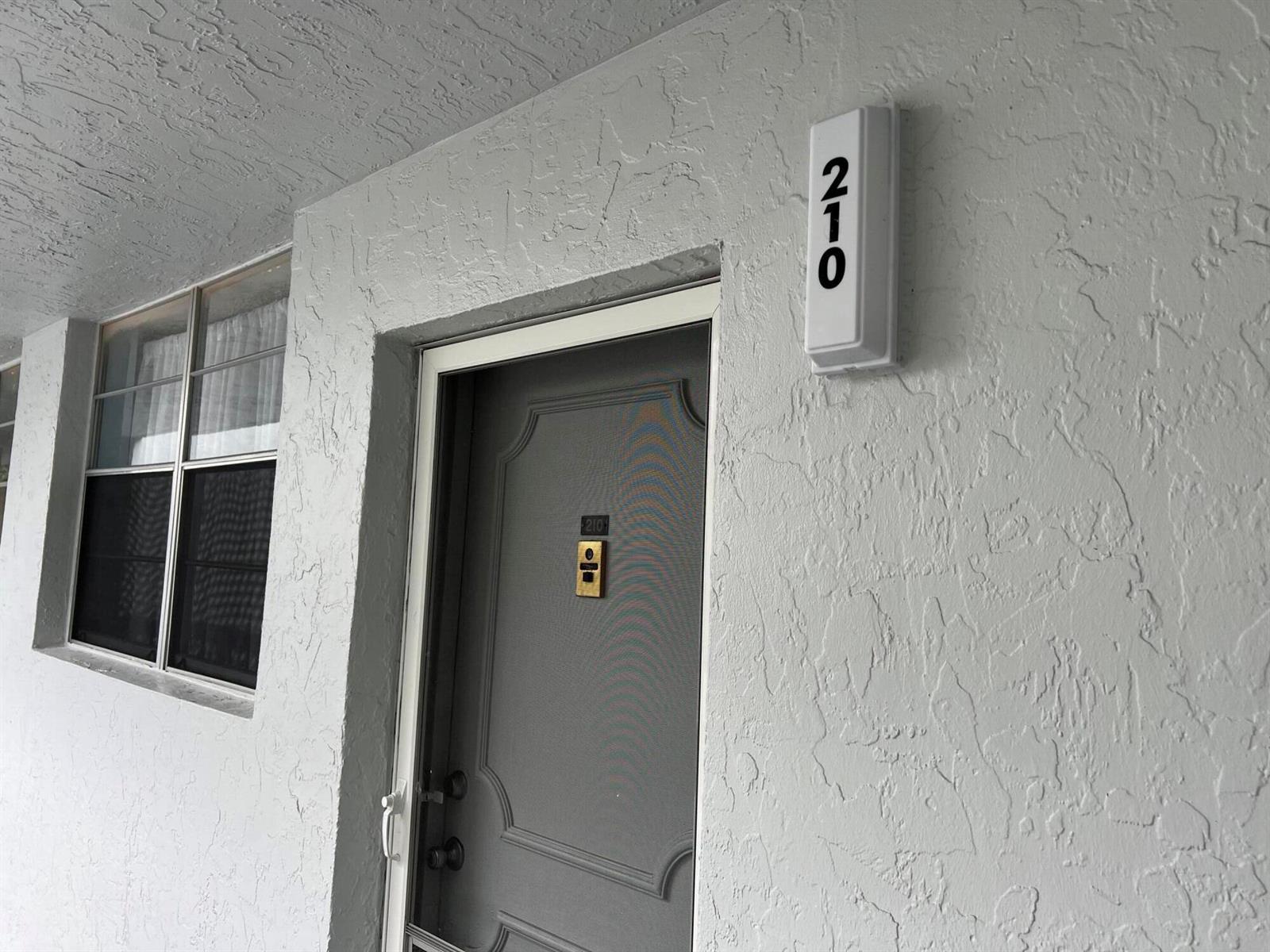 A gray apartment door with a screen door and the number 210 displayed above — 3220 Holiday Springs Boulevard # 210, Margate FL 33063, MARGATE, FL