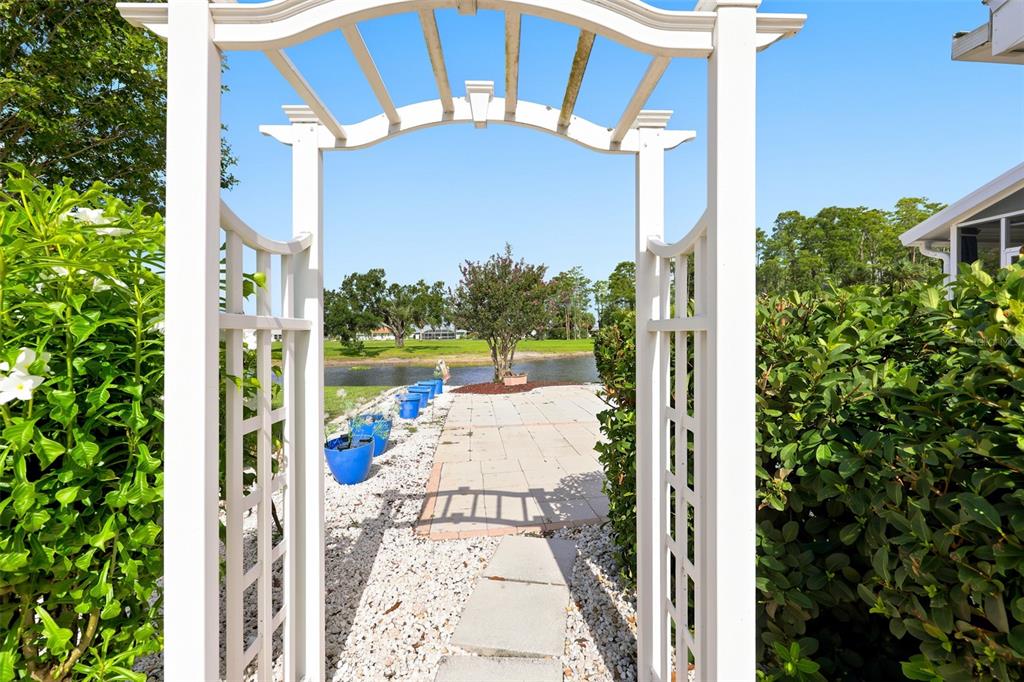 A white garden arbor frames a stone patio overlooking a canal and green lawn — 1002 EL RANCHO DRIVE, SUN CITY CENTER, FL