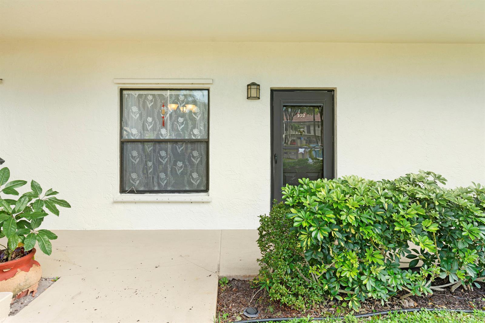 A white exterior wall features a window with patterned curtains and a dark front door — 3 Vía De Casas Sur # 102, Boynton Beach FL 33426, BOYNTON BEACH, FL