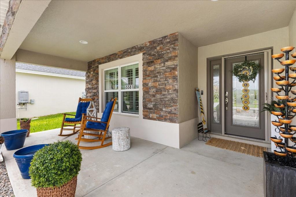 A covered front porch features stone veneer siding, two wooden rocking chairs, and a front door — 5581 PASSION FLOWER WAY, THE VILLAGES, FL