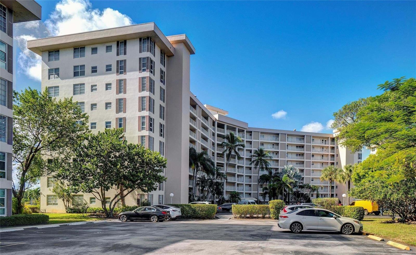A multi-story apartment building with a parking lot in the foreground and surrounding trees — 2681 S Course Drive # 712, Pompano Beach FL 33069, POMPANO BEACH, FL