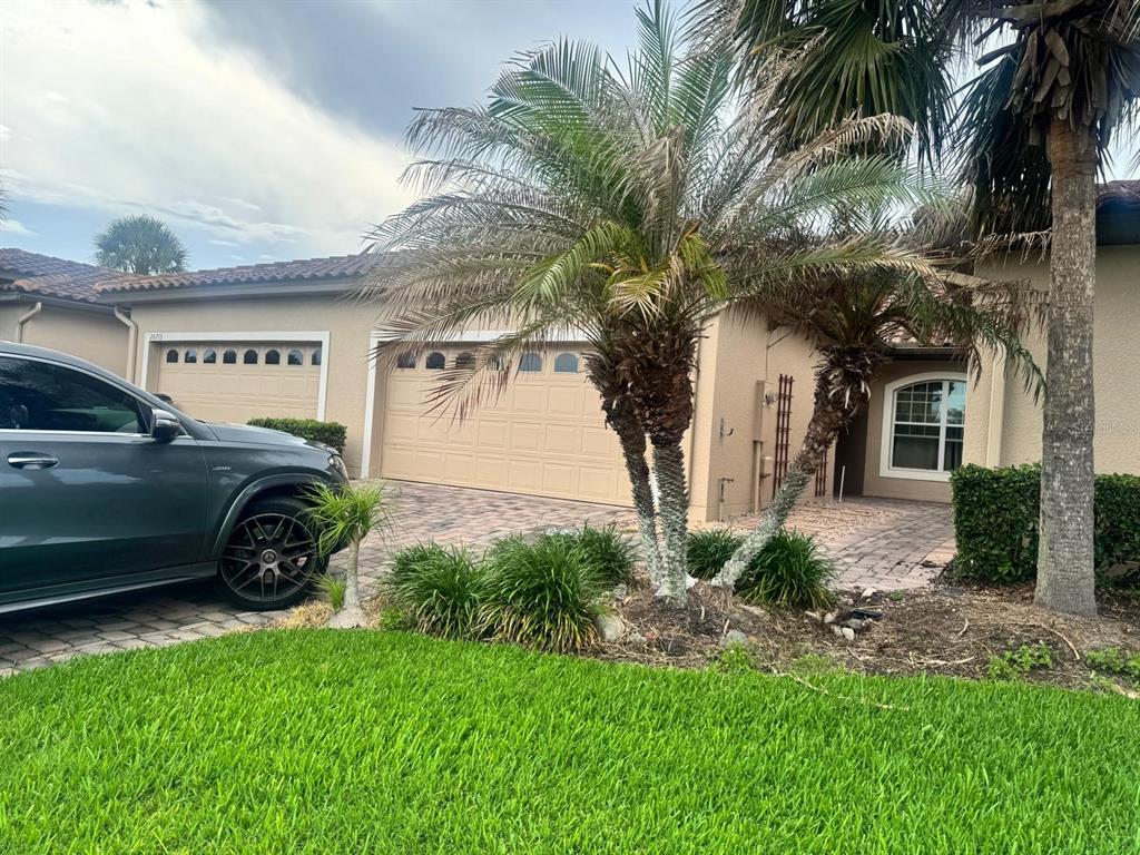 A beige house exterior features a paver driveway, two garage doors, and several palm trees — 26715 BELLA VISTA DRIVE, HOWEY IN THE HILLS, FL
