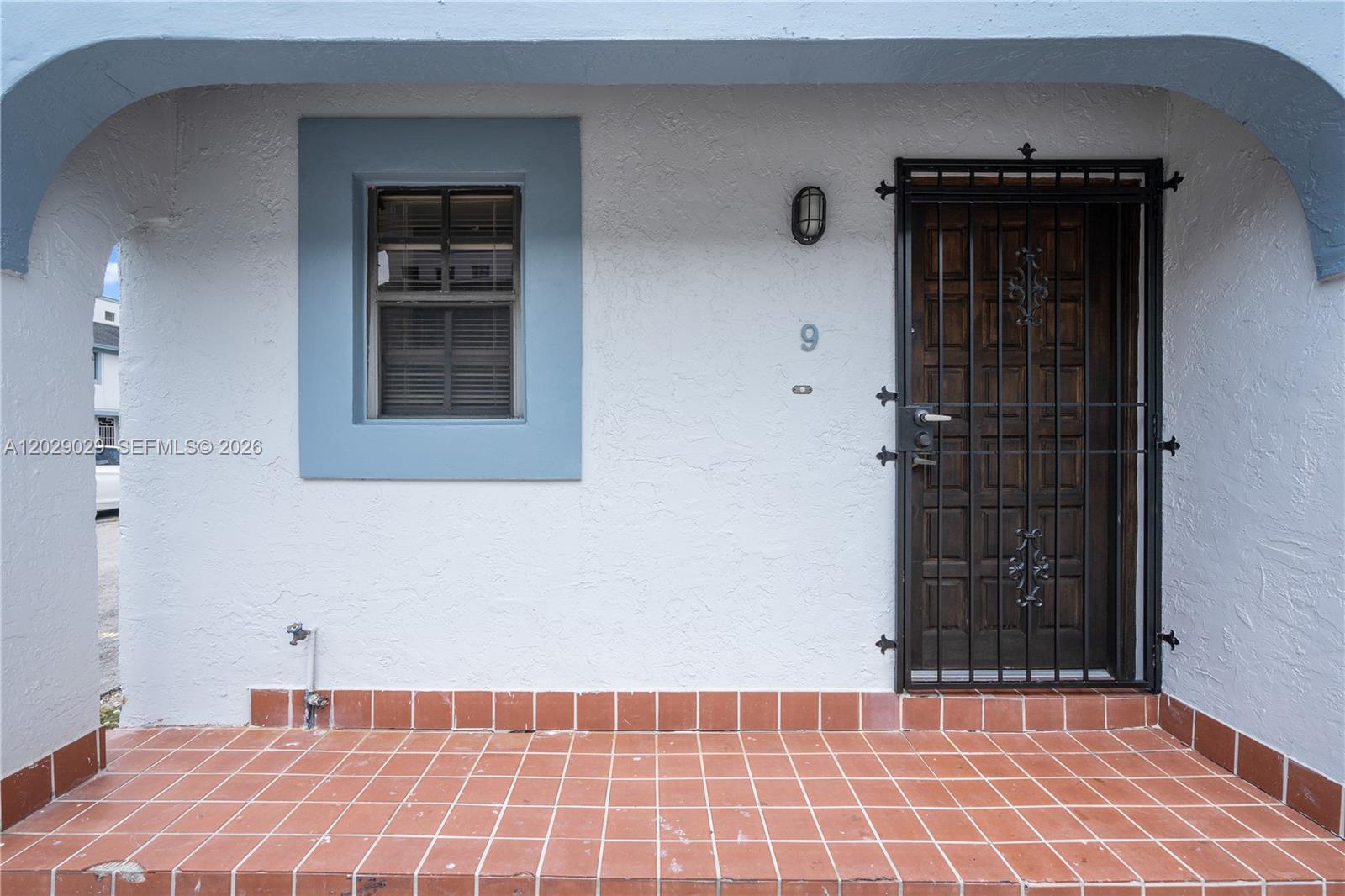 A white exterior wall features a window with blue trim, a door with a security gate, and tiled flooring — 12035 SW 18th St # 48, Miami FL 33175, MIAMI, FL