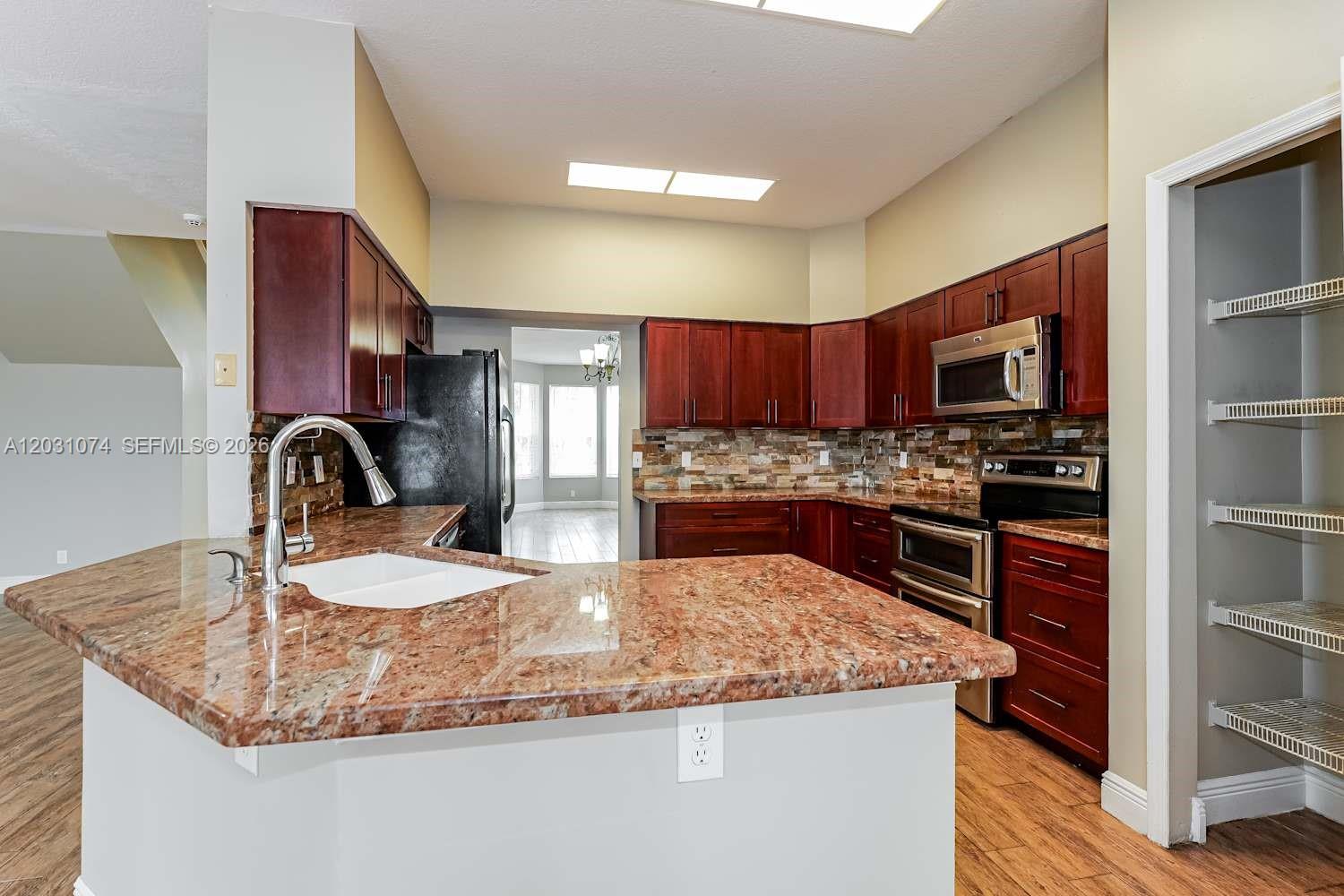 A kitchen features dark wood cabinets, granite countertops, a stone backsplash, and stainless steel appliances — WELLINGTON, FL