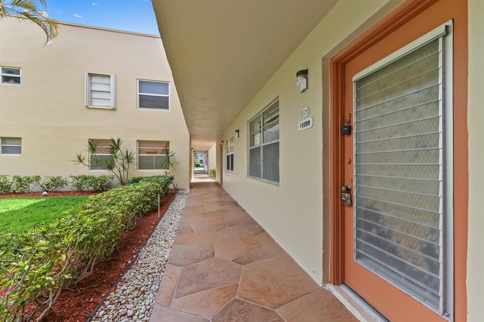 An outdoor walkway leads past a tan apartment building with a numbered orange door — 211 Piedmont E, Delray Beach FL 33484, DELRAY BEACH, FL