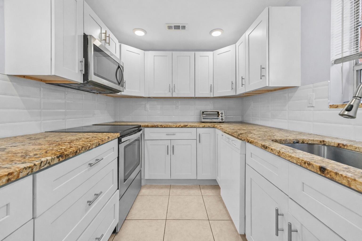 A galley kitchen features white cabinets, granite countertops, a stainless steel microwave, and tile flooring — 211 Piedmont E, Delray Beach FL 33484, DELRAY BEACH, FL