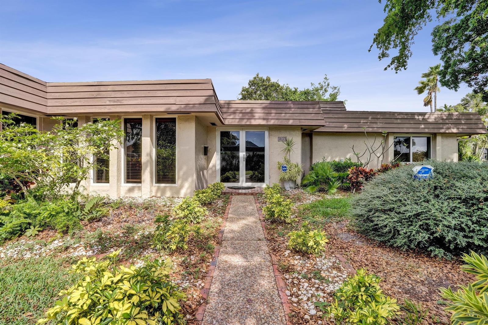 A single-story house with a flat roof, beige stucco walls, and a central glass-paned entryway — 6207 Jacaranda Circle, Tamarac FL 33319, TAMARAC, FL