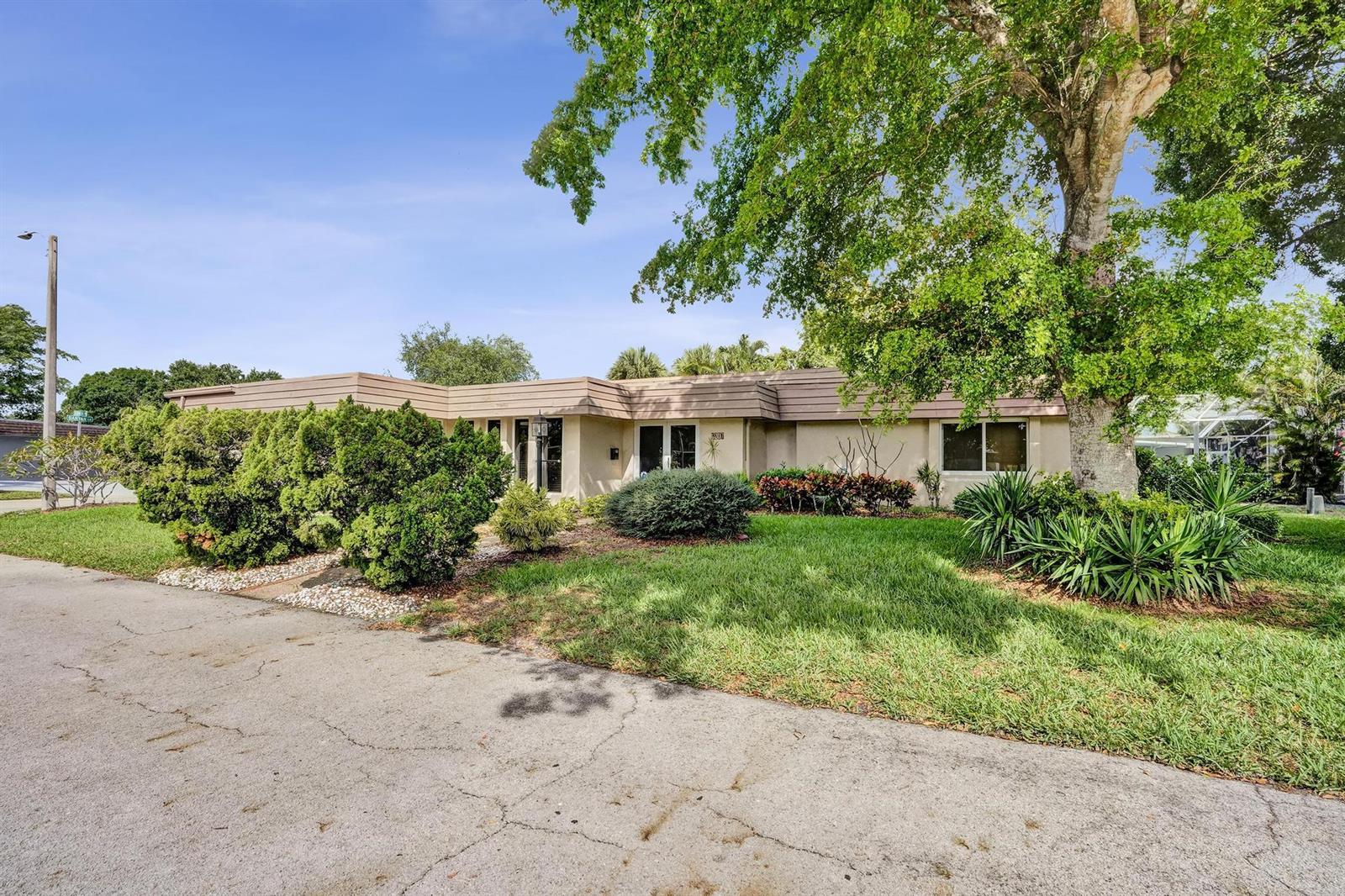 A single-story house with a flat roof, tan exterior, and mature landscaping under a blue sky — 6207 Jacaranda Circle, Tamarac FL 33319, TAMARAC, FL