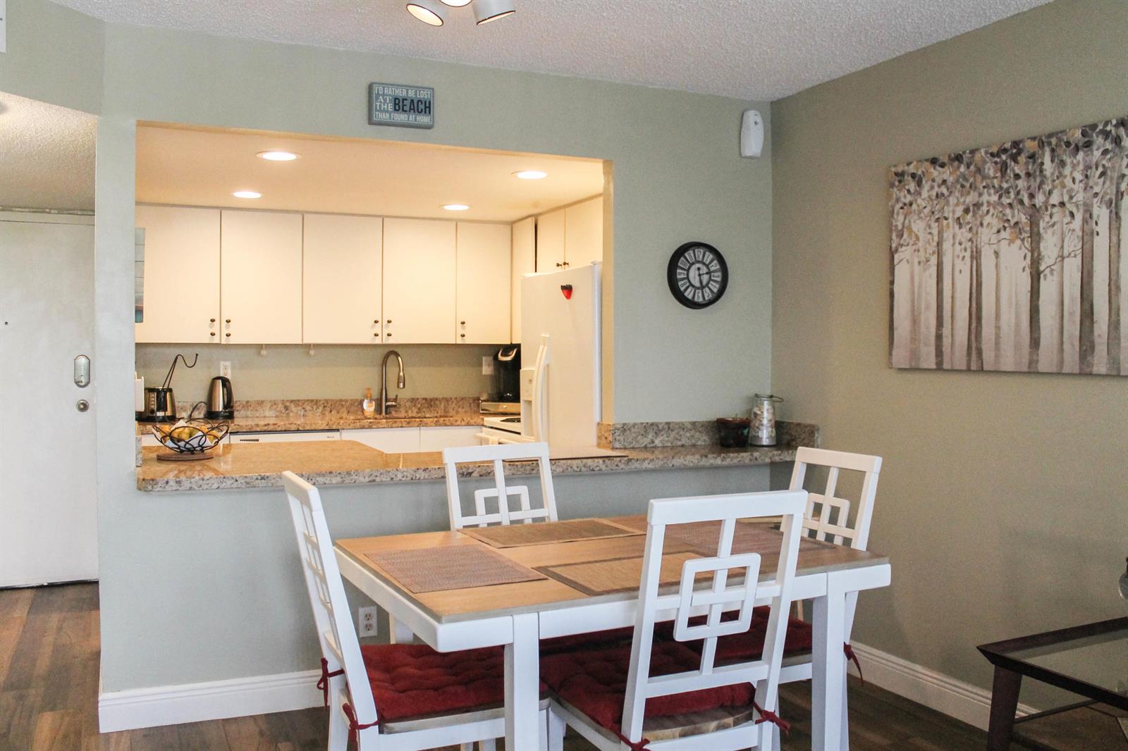A dining area with a white table and chairs sits before an open kitchen pass-through — 1830 Embassy Drive # 306, West Palm Beach FL 33401, WEST PALM BEACH, FL