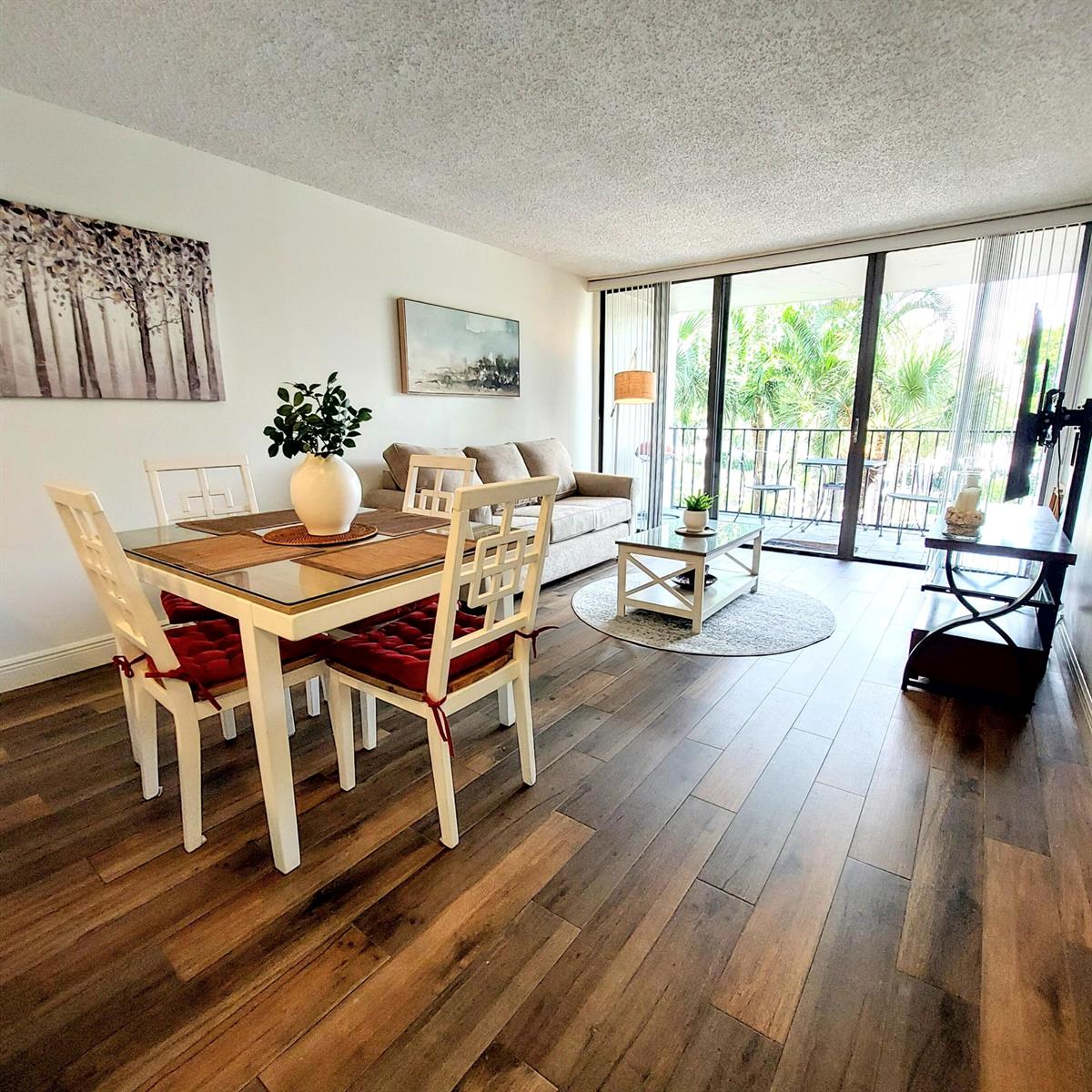 A living and dining area with wood-look flooring, white furniture, and a balcony with greenery — 1830 Embassy Drive # 306, West Palm Beach FL 33401, WEST PALM BEACH, FL