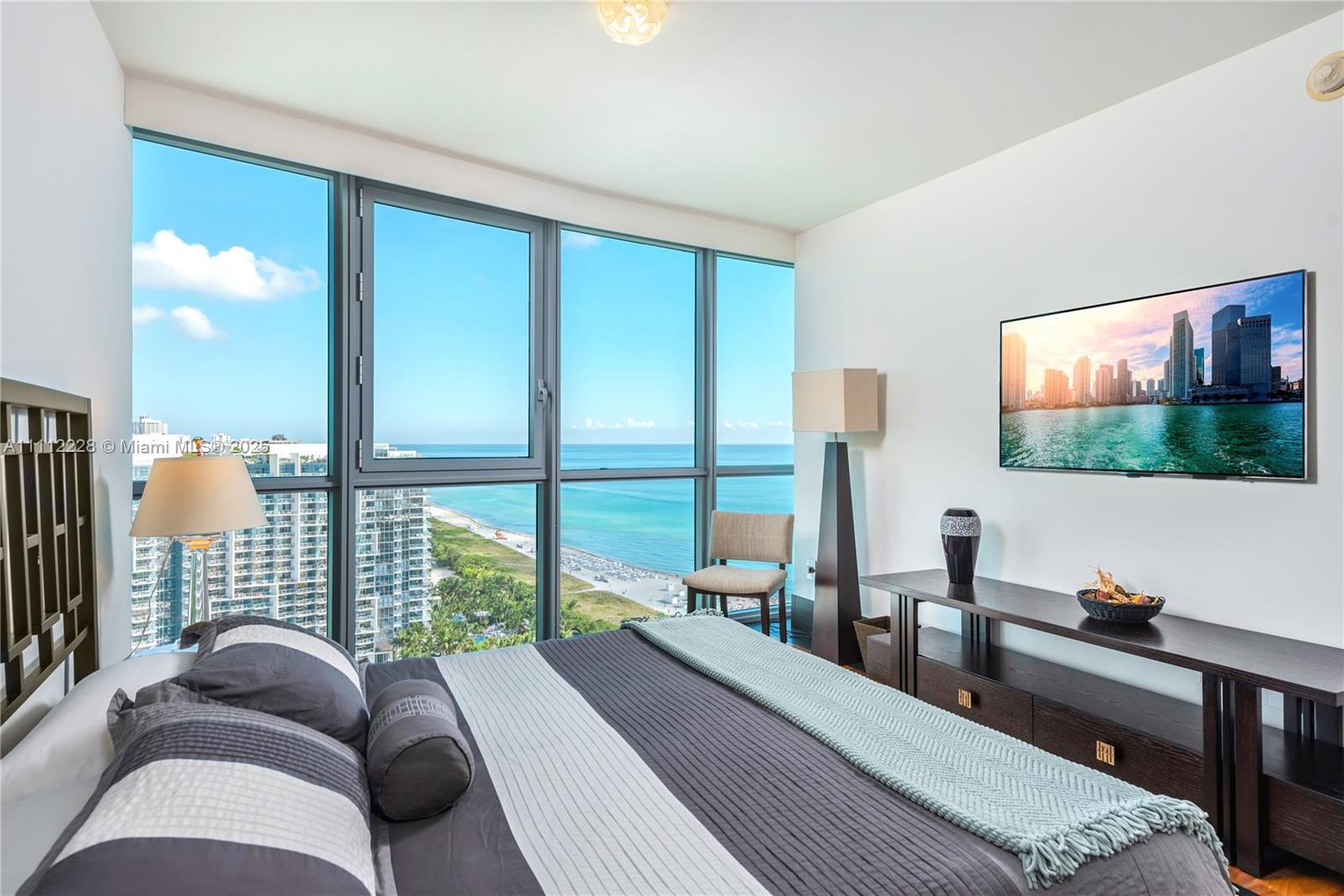 A bedroom features a bed, a wall-mounted television, and floor-to-ceiling windows overlooking the ocean — 101 20th St # 2707, Miami Beach FL 33139, MIAMI BEACH, FL