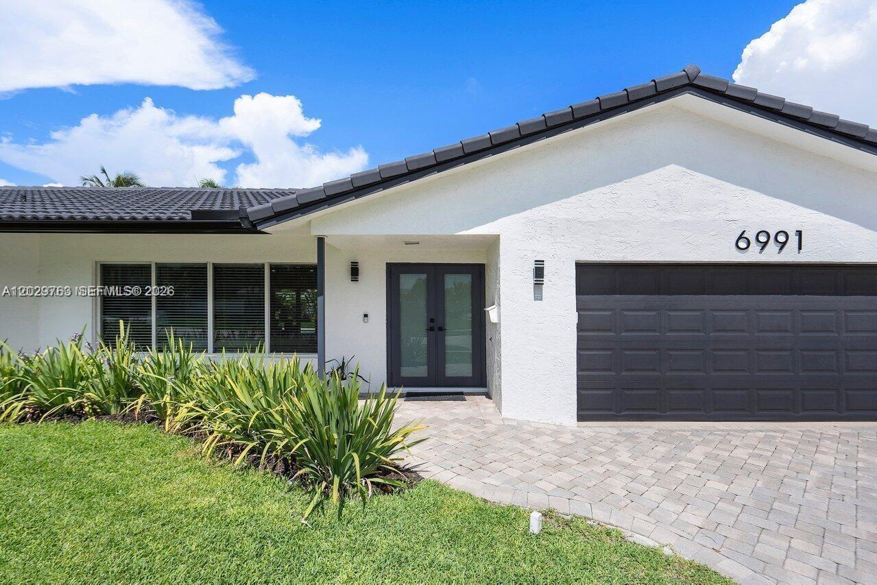 A white house exterior features a dark garage door, double front doors, and a paved driveway — 6991 NE 7th Ave # 0, Boca Raton FL 33487, BOCA RATON, FL