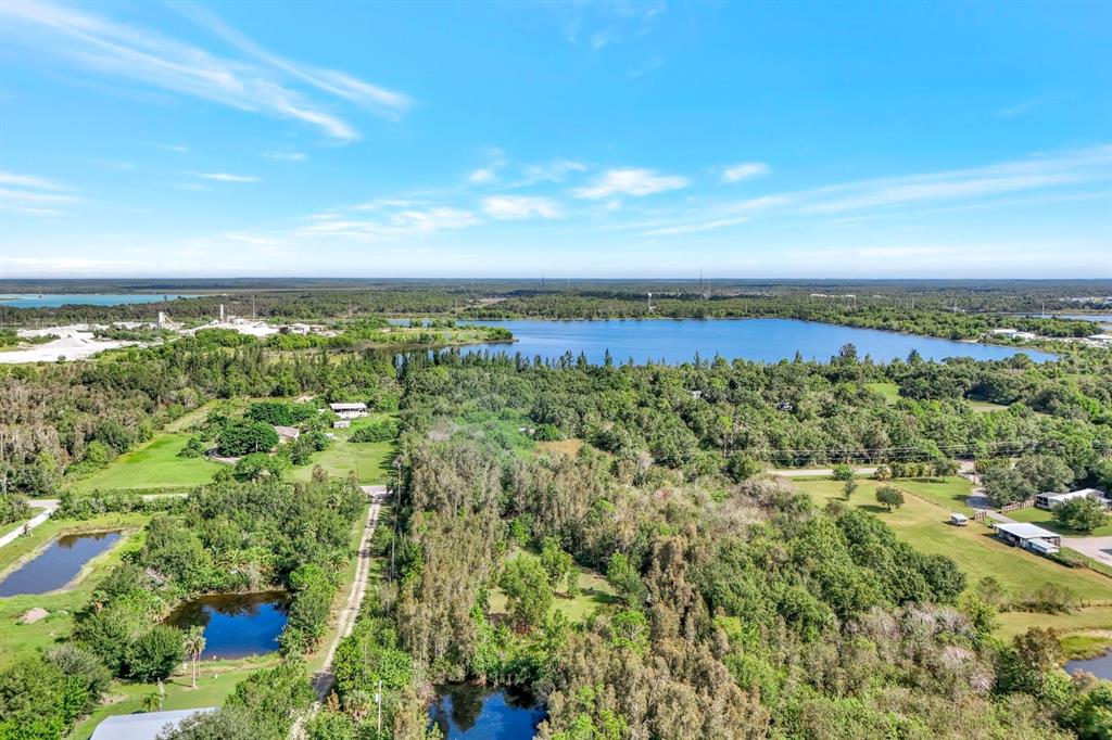 An aerial view shows a large lake surrounded by dense trees, grassy lots, and small buildings — 41601 LITTLE FARM ROAD, PUNTA GORDA, FL