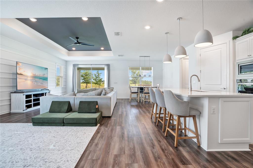 An open-concept living room and kitchen feature a tray ceiling, wood-look flooring, and island seating — 11237 ROLLING FORK TRAIL, PARRISH, FL