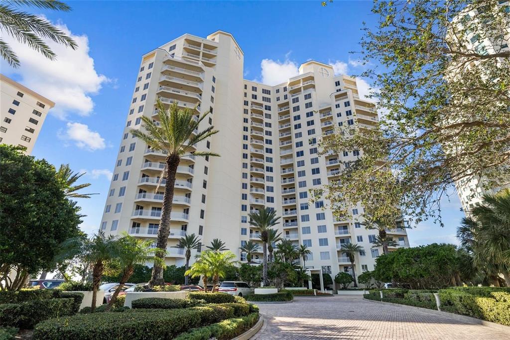 A tall, multi-story beige residential building stands behind a paved driveway and landscaped gardens — 1200 GULF BOULEVARD UNIT 1001, CLEARWATER, FL