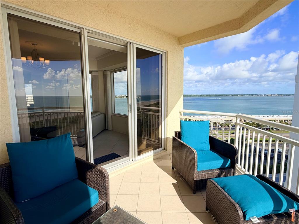 A balcony with wicker chairs and blue cushions overlooks a coastal view through glass doors — 1200 GULF BOULEVARD UNIT 1001, CLEARWATER, FL