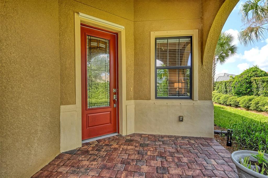 A red front door with a glass insert sits beside a window on a paved porch — 6743 HAVERHILL COURT, BRADENTON, FL
