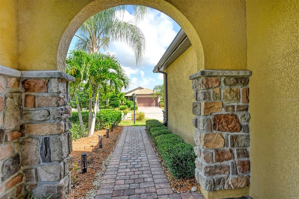 A stone-pillared archway frames a brick walkway leading toward a house with a garage — 6743 HAVERHILL COURT, BRADENTON, FL