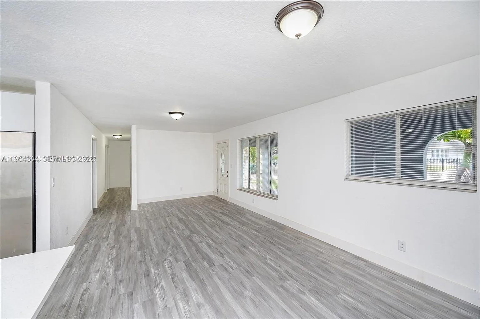 An empty living room features white walls, gray wood-look flooring, and two windows with blinds — 11950 SW 182nd Ter, Miami FL 33177, MIAMI, FL