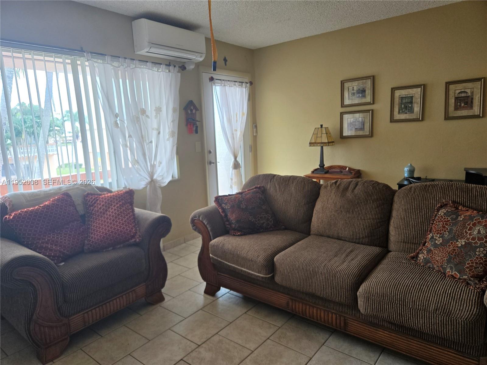 A living room features a brown sofa and loveseat, tile flooring, and a wall-mounted air conditioner — 900 SW 11th Ave # 12A, Hallandale Beach FL 33009, HALLANDALE BEACH, FL