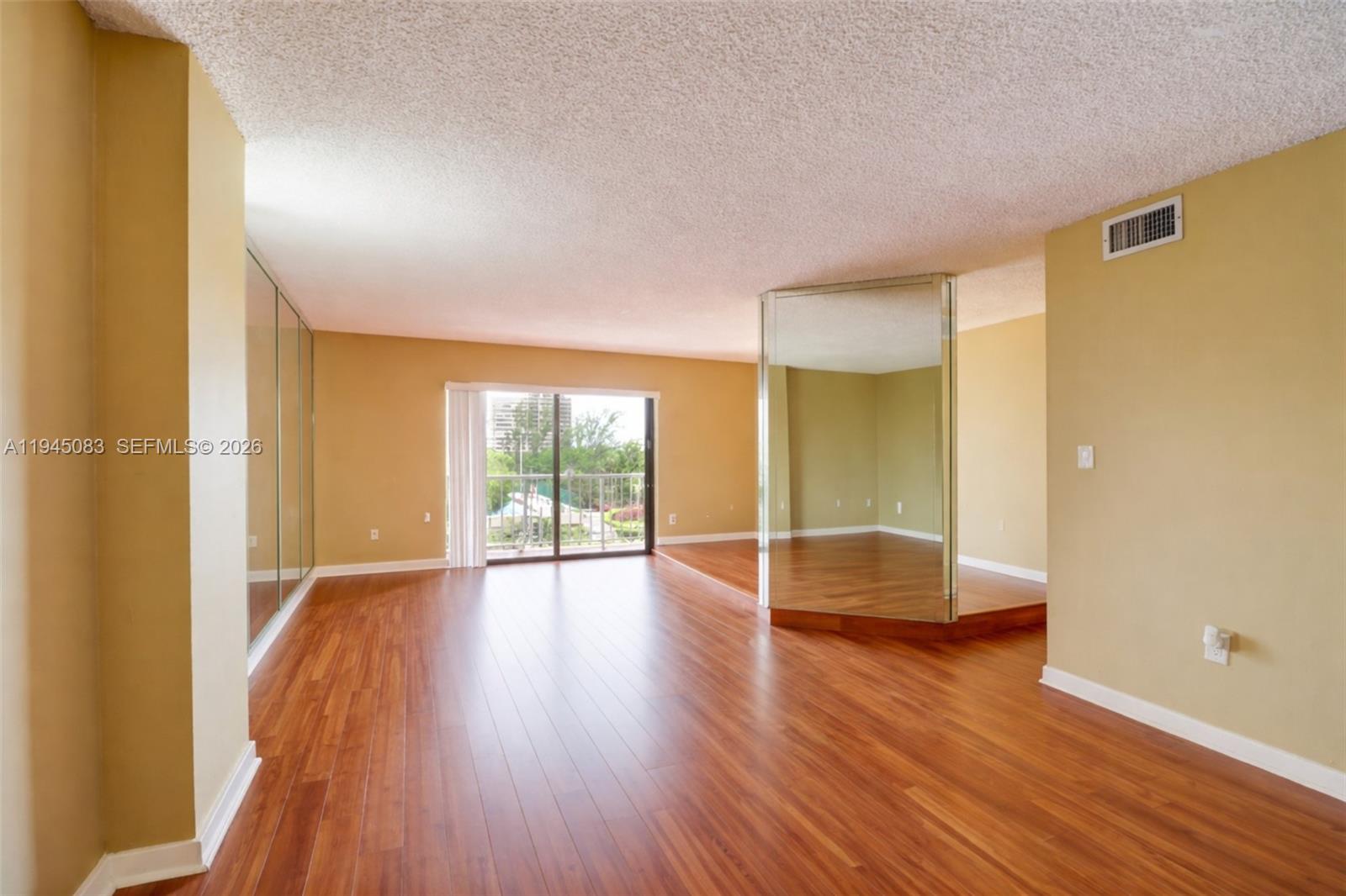 An empty room features hardwood floors, mirrored walls, and a sliding glass door to a balcony — 1650 NE 115th St # 505, Miami FL 33181, MIAMI, FL