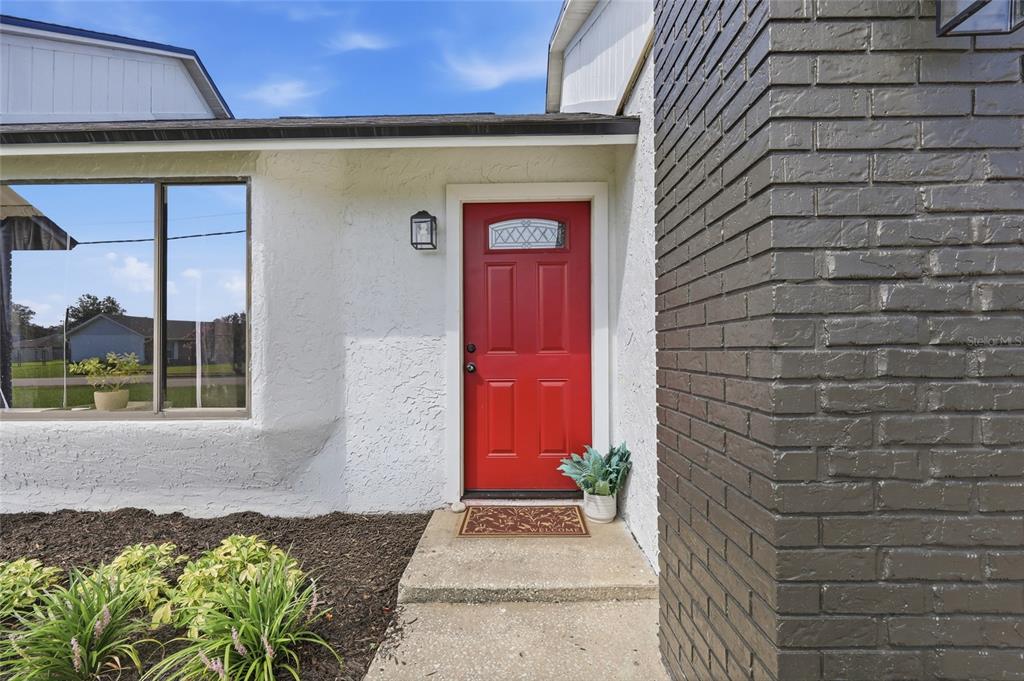 A red front door is set in a white stucco wall beside a brick chimney — 2643 SALTERS COURT, DELTONA, FL