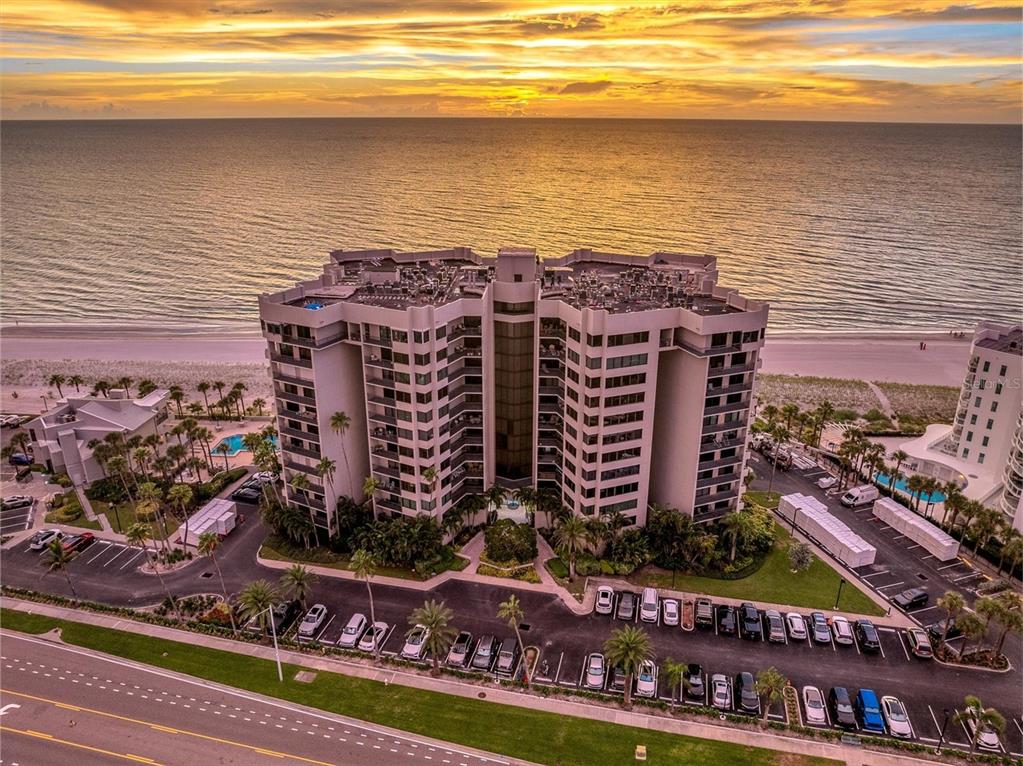 An aerial view shows a multi-story beachfront condominium building with a parking lot at sunset — 1600 GULF BOULEVARD UNIT 617, CLEARWATER BEACH, FL