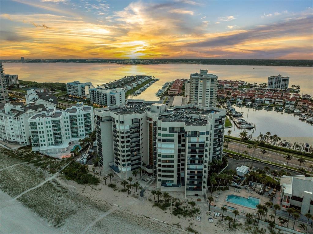 An aerial view shows beachfront high-rise condominiums, a marina, and a sunset over the water — 1600 GULF BOULEVARD UNIT 617, CLEARWATER BEACH, FL