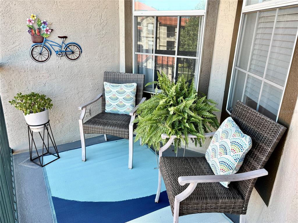 A balcony features two wicker chairs with patterned pillows, a potted plant, and wall decor — 18107 PEREGRINES PERCH PLACE UNIT 6303, LUTZ, FL