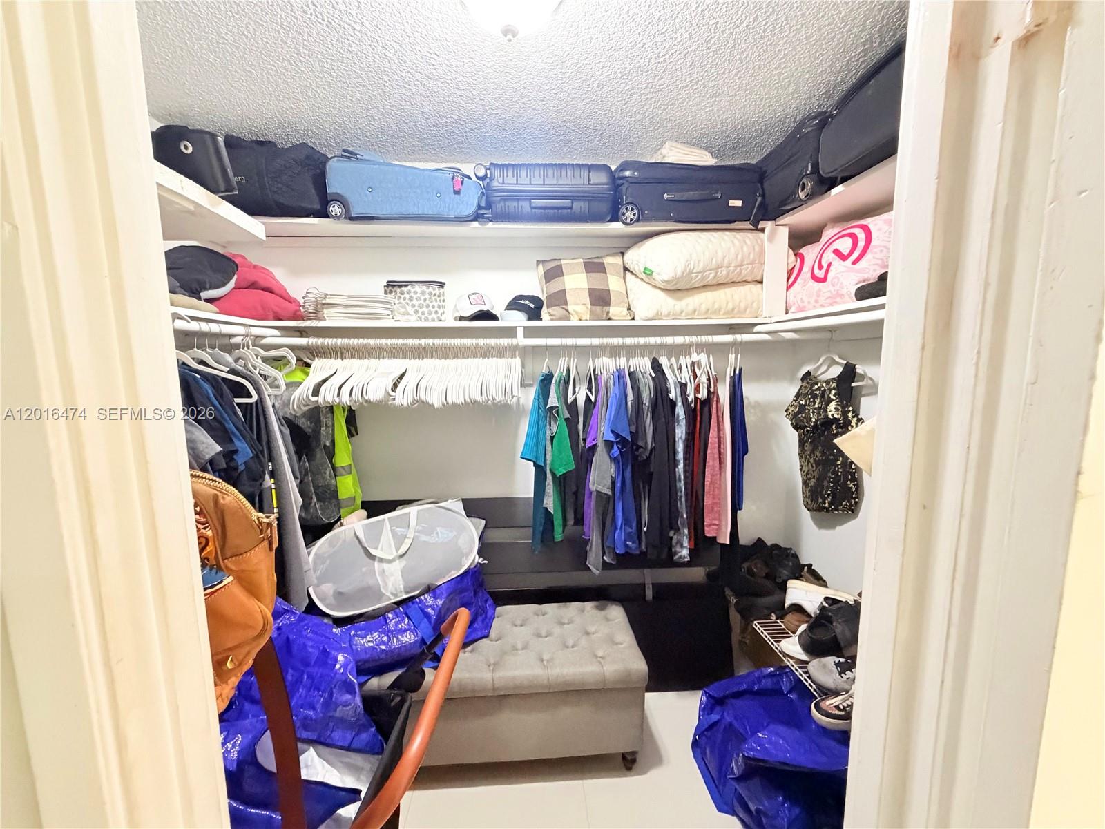 A walk-in closet features white shelving, hanging clothes, luggage, and a tufted storage ottoman — 1680 NE 191st St # 2, Miami FL 33179, MIAMI, FL