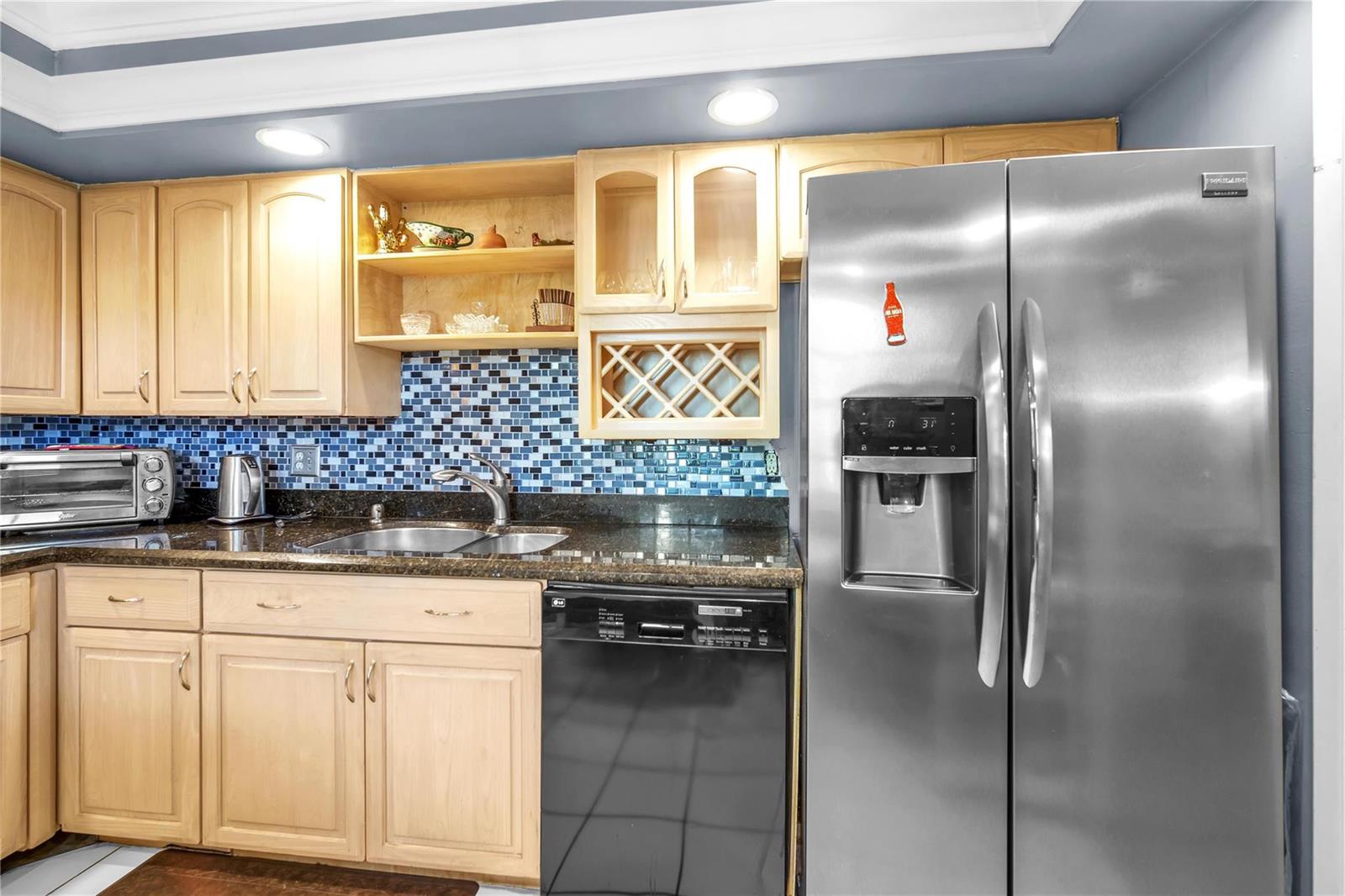 A kitchen with light wood cabinets, blue mosaic tile backsplash, dark countertops, and stainless steel appliances — 400 Oaks Lane # 115, Pompano Beach FL 33069, POMPANO BEACH, FL