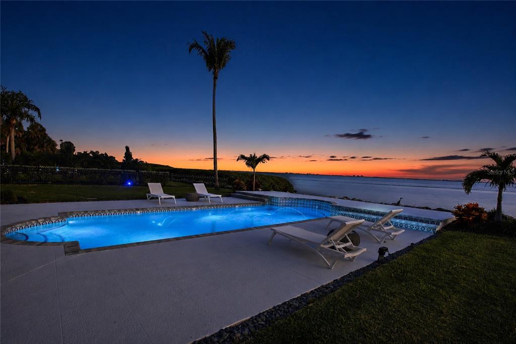 An illuminated swimming pool sits on a patio overlooking a body of water at sunset — 7524 WESTMORELAND DRIVE, SARASOTA, FL