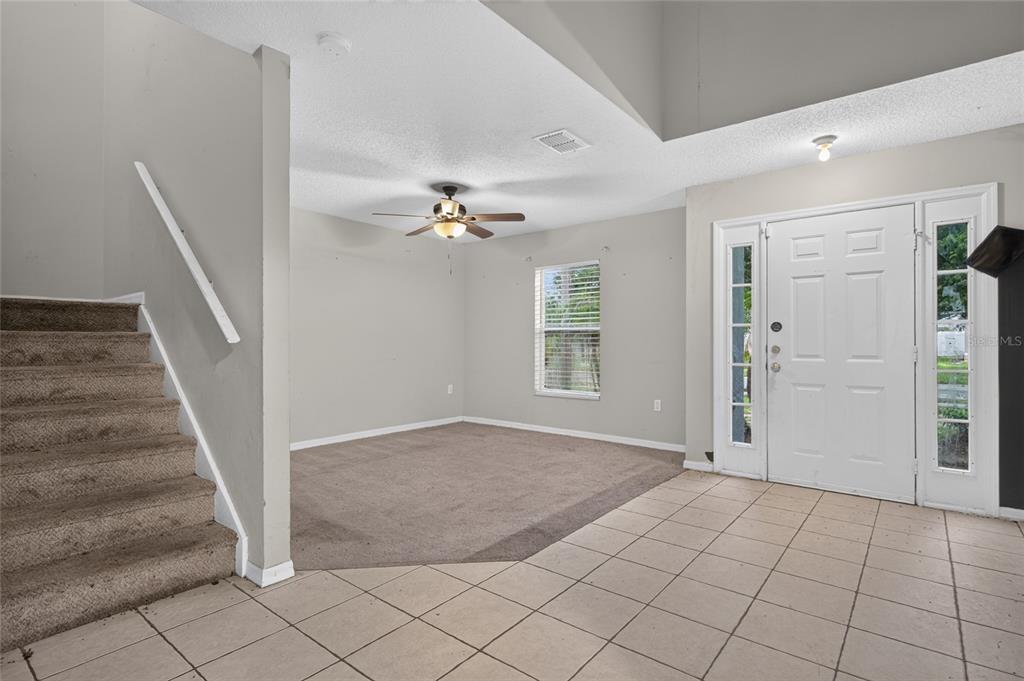A carpeted living room with a ceiling fan sits adjacent to a tiled entryway and staircase — 12803 LAKE VISTA DRIVE, GIBSONTON, FL