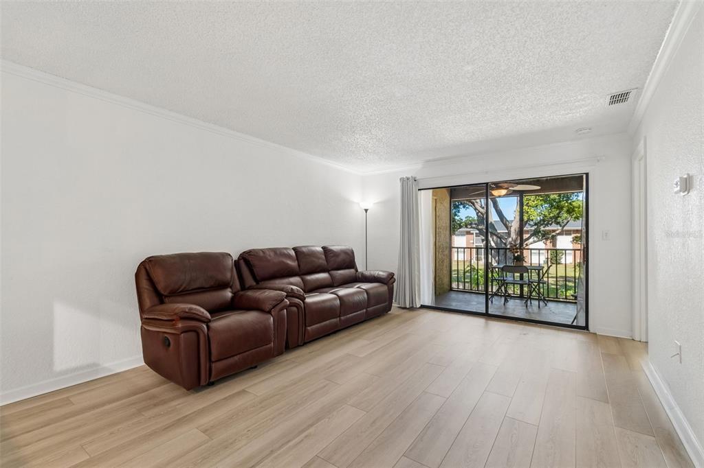 A living room features light wood-look flooring, a brown leather sofa, and a sliding glass door — 10100 SAILWINDS BOULEVARD N UNIT 202, LARGO, FL