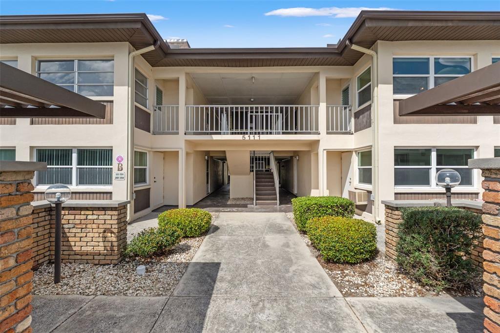 A two-story apartment building exterior features a central staircase, concrete walkway, and landscaped bushes — 5111 AMULET DRIVE UNIT 202, NEW PORT RICHEY, FL