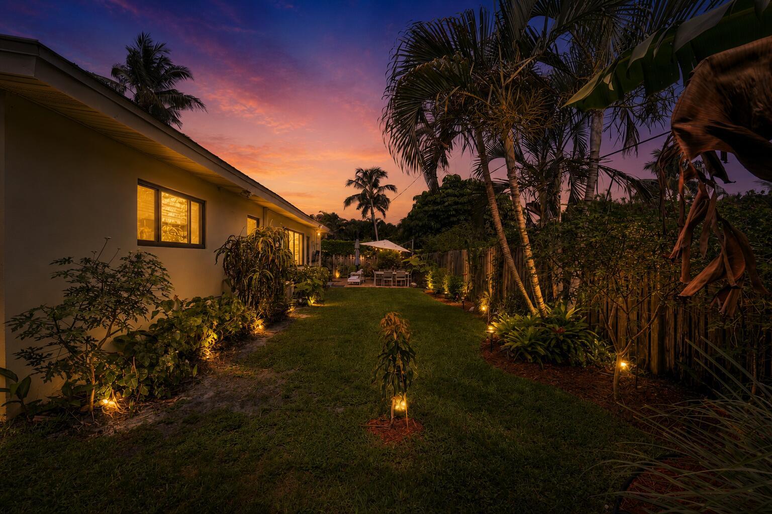 A backyard lawn at sunset features landscape lighting, palm trees, and a patio dining area — 109 NE 17th Street, Delray Beach FL 33444, DELRAY BEACH, FL