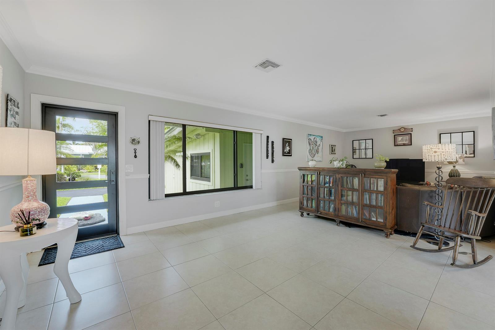 A bright living room features light tile flooring, a glass-paneled front door, and a wooden bookcase — 109 NE 17th Street, Delray Beach FL 33444, DELRAY BEACH, FL