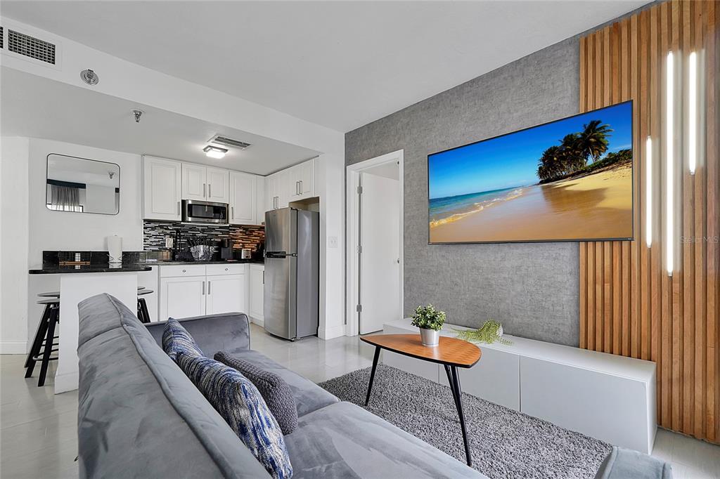 A living room with a grey sofa, a wall-mounted television, and an adjacent open-concept kitchen — 6165 CARRIER DRIVE UNIT 3701, ORLANDO, FL
