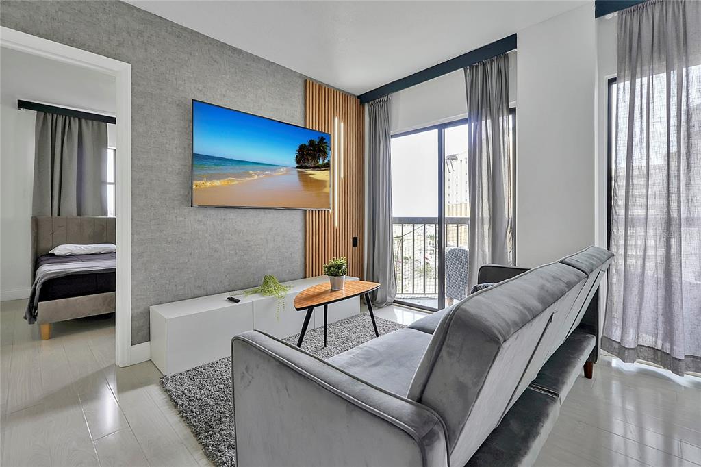 A living room features a wall-mounted television, wood slat accent, grey sofa, and balcony access — 6165 CARRIER DRIVE UNIT 3701, ORLANDO, FL