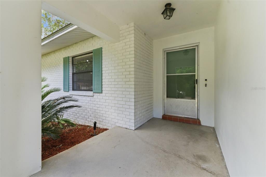 A covered concrete porch leads to a white brick exterior wall with a green-shuttered window — 8127 SW 12TH PLACE, GAINESVILLE, FL