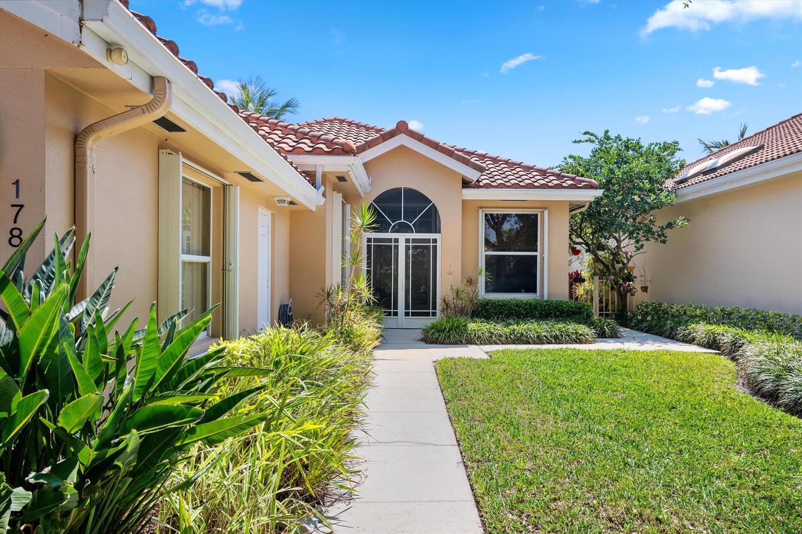 A beige single-story house features a tiled roof, a front walkway, and landscaped garden beds — 178 Hampton Place, Jupiter FL 33458, JUPITER, FL